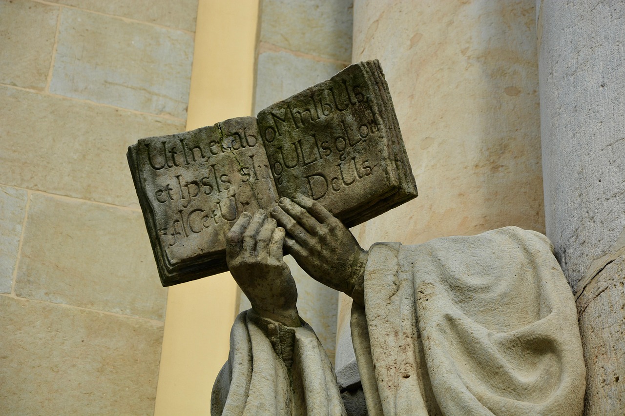 fej nélküli kőszobor, kezében a néző felé fordított, latin nyelvű Biblia
